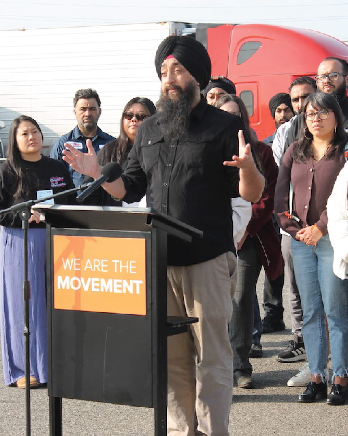 A man wearing a black turban and black shirt speaks passionately at a black podium outdoors, gesturing with his hands. The podium has an orange sign that reads 