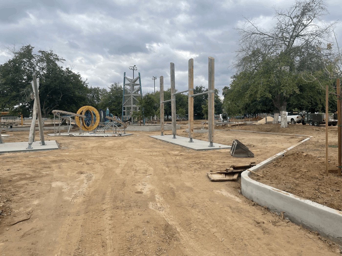 A wide landscape view of the park's construction site under a cloudy sky. A newly poured concrete pathway curves through the bare earth in the foreground. In the background, various playground installations are visible, including tall upright wooden logs, the circular rope climbing structure, and the tall wooden tower.