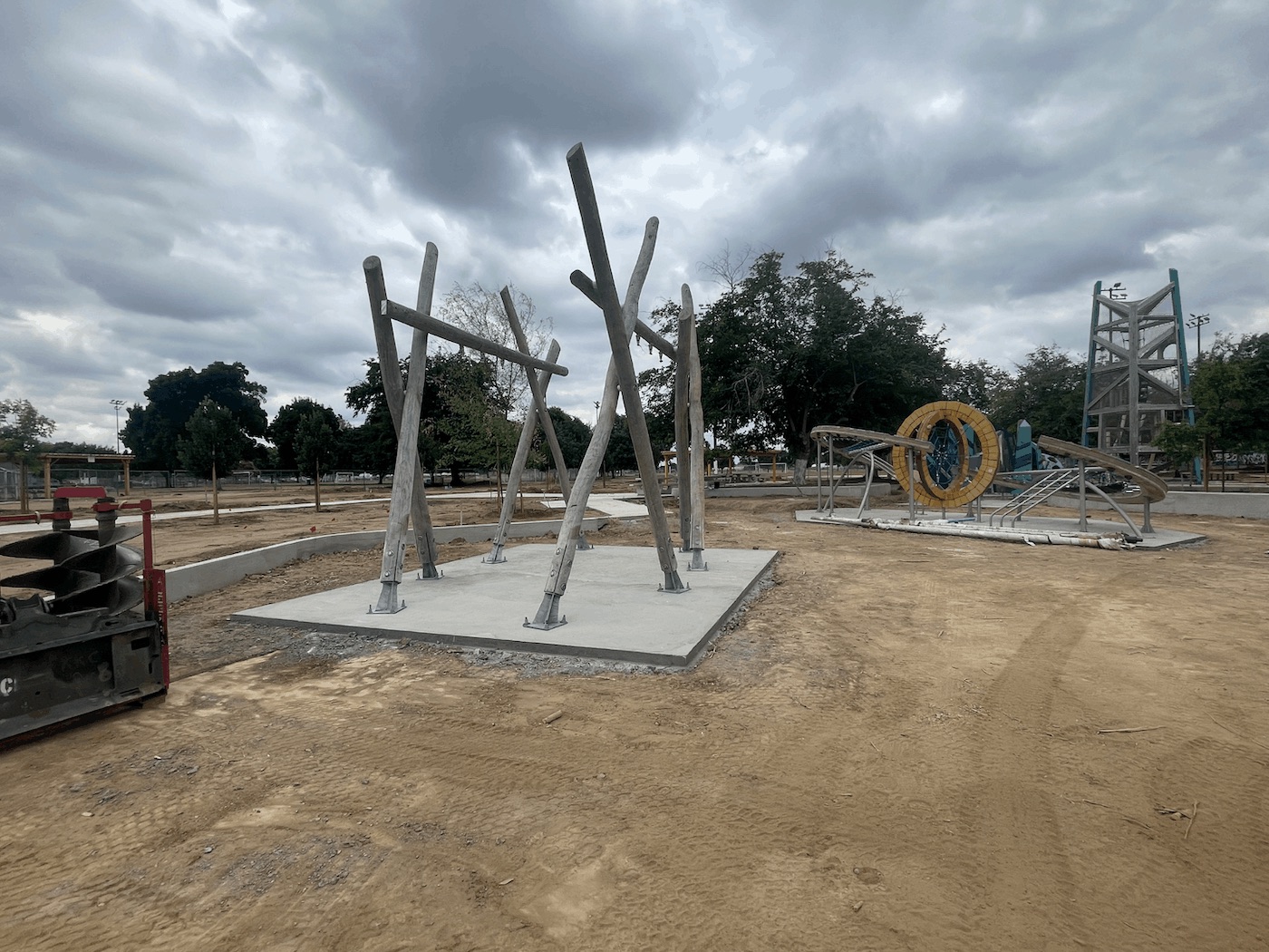 A minimalist playground structure consisting of tall, thick wooden logs leaning together to support horizontal bars with hanging rings. The logs are secured to a square concrete pad with heavy metal brackets. The surrounding park area consists of graded dirt, with other playground equipment visible in the background.