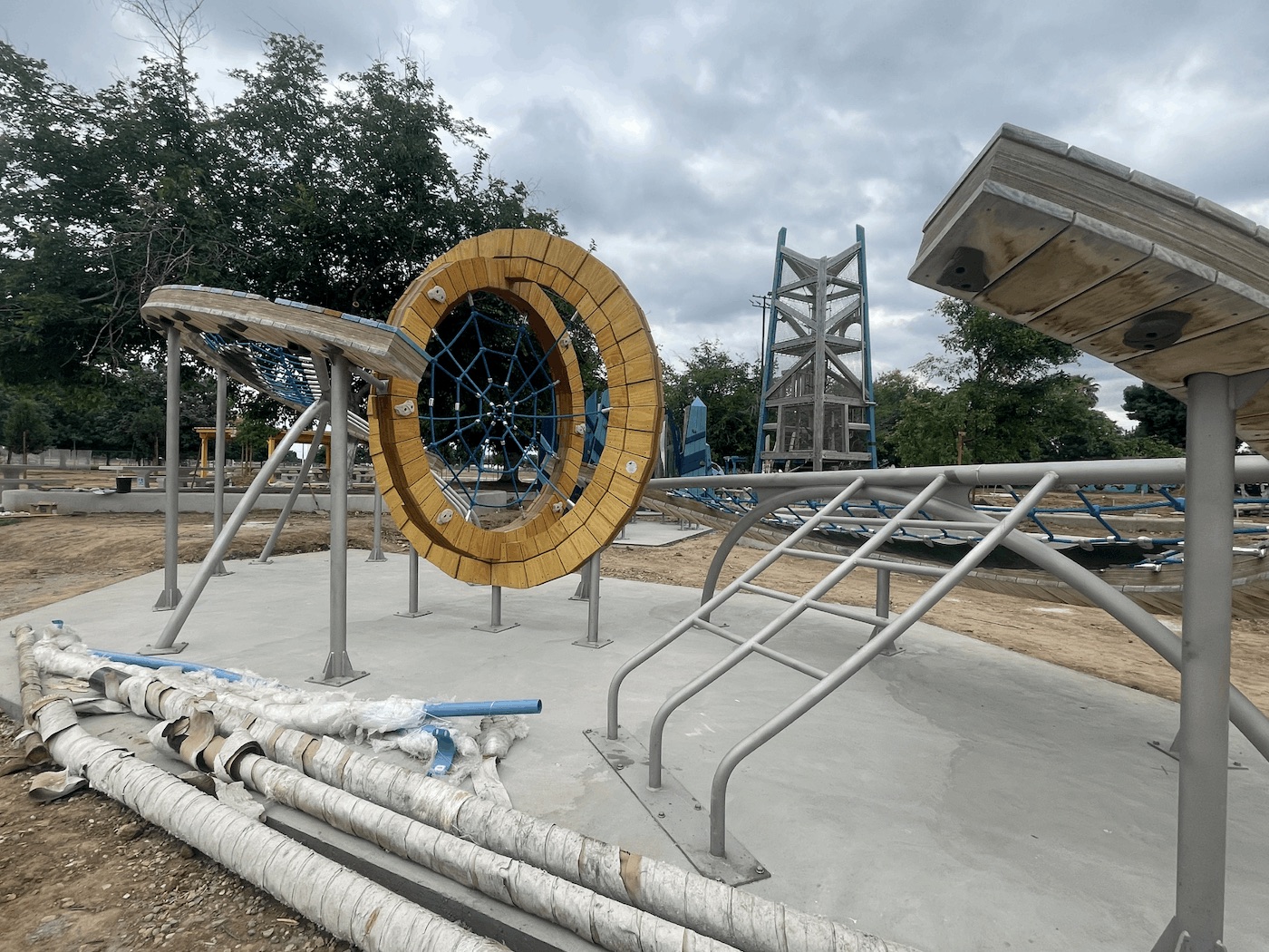 A wide angle of a complex playground structure featuring two large, upright wooden rings framing a blue rope spiderweb climbing net. Connected to the rings are curved metal bars, a rope hammock, and sloped wooden platforms. The equipment sits on concrete amidst the unfinished dirt landscaping of the park.