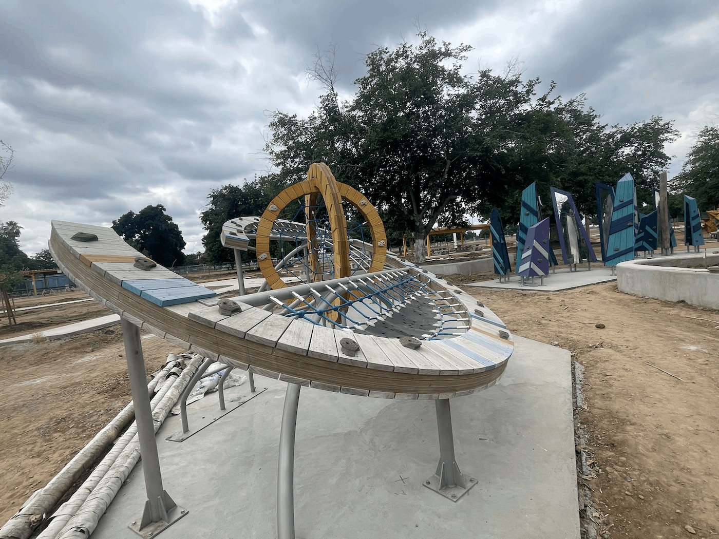 A unique, modern playground structure made of a curved metal and wood frame with a grid of blue ropes strung across it, creating a suspended hammock or climbing net. The structure sits on a concrete pad surrounded by a dirt construction site, with circular climbing elements visible in the background.