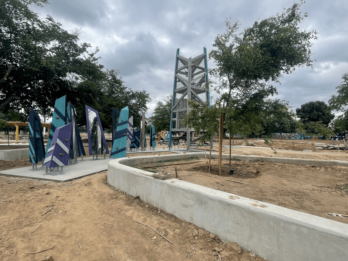 A perspective shot of the park's new playground area showing the angled blue and teal climbing walls on the left and the tall wooden play tower in the center. In the foreground, newly planted saplings supported by wooden stakes sit inside curved concrete planter walls filled with dirt.