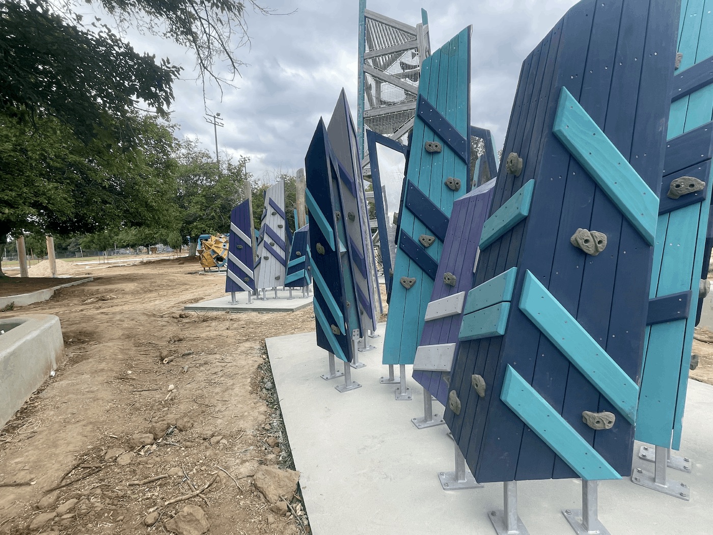 A close-up ground-level view of several angled, freestanding climbing walls in a park construction site. The walls are constructed of wood painted in shades of blue, teal, and gray, and feature attached rock-climbing holds. They are mounted on silver metal posts secured to a new concrete surface.
