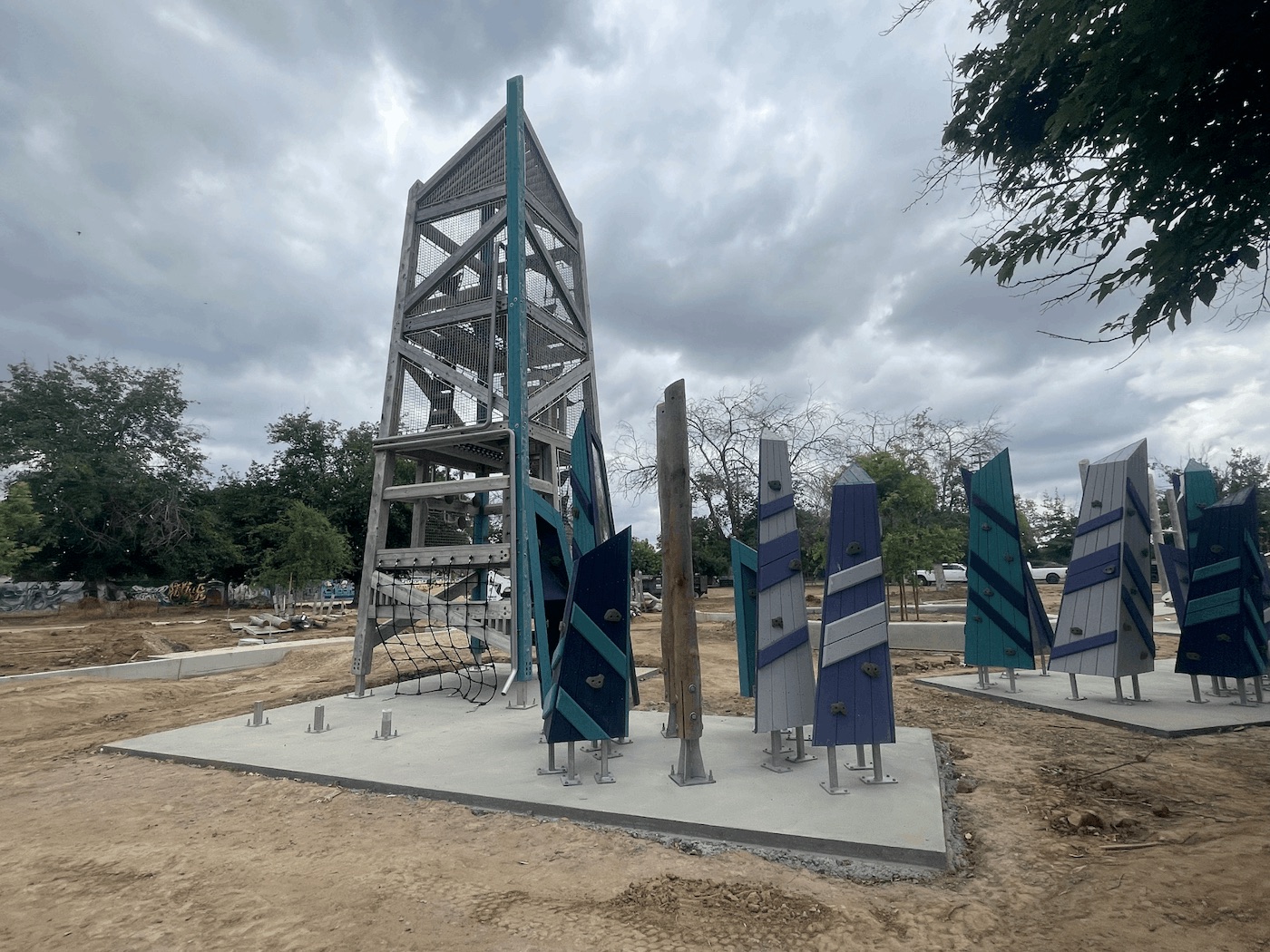 A tall, modern wooden playground tower with teal metal accents and interior safety netting, standing next to a cluster of freestanding, angled climbing walls. The climbing walls are painted in geometric patterns of blue, teal, purple, and gray. The equipment sits on a fresh concrete pad surrounded by dirt.