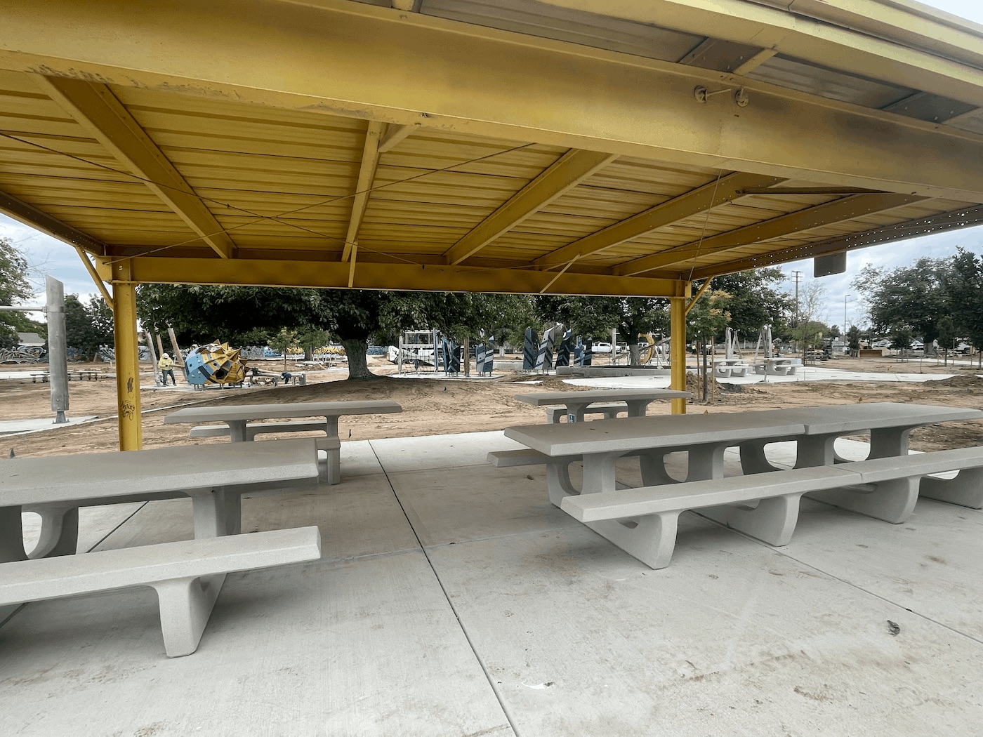 A view from underneath a large yellow metal picnic shelter, looking out over a park under construction. Several heavy concrete picnic tables sit on a finished concrete pad in the foreground. In the background, modern playground structures, including a tall tower and climbing walls, are visible amidst bare dirt and scattered trees.