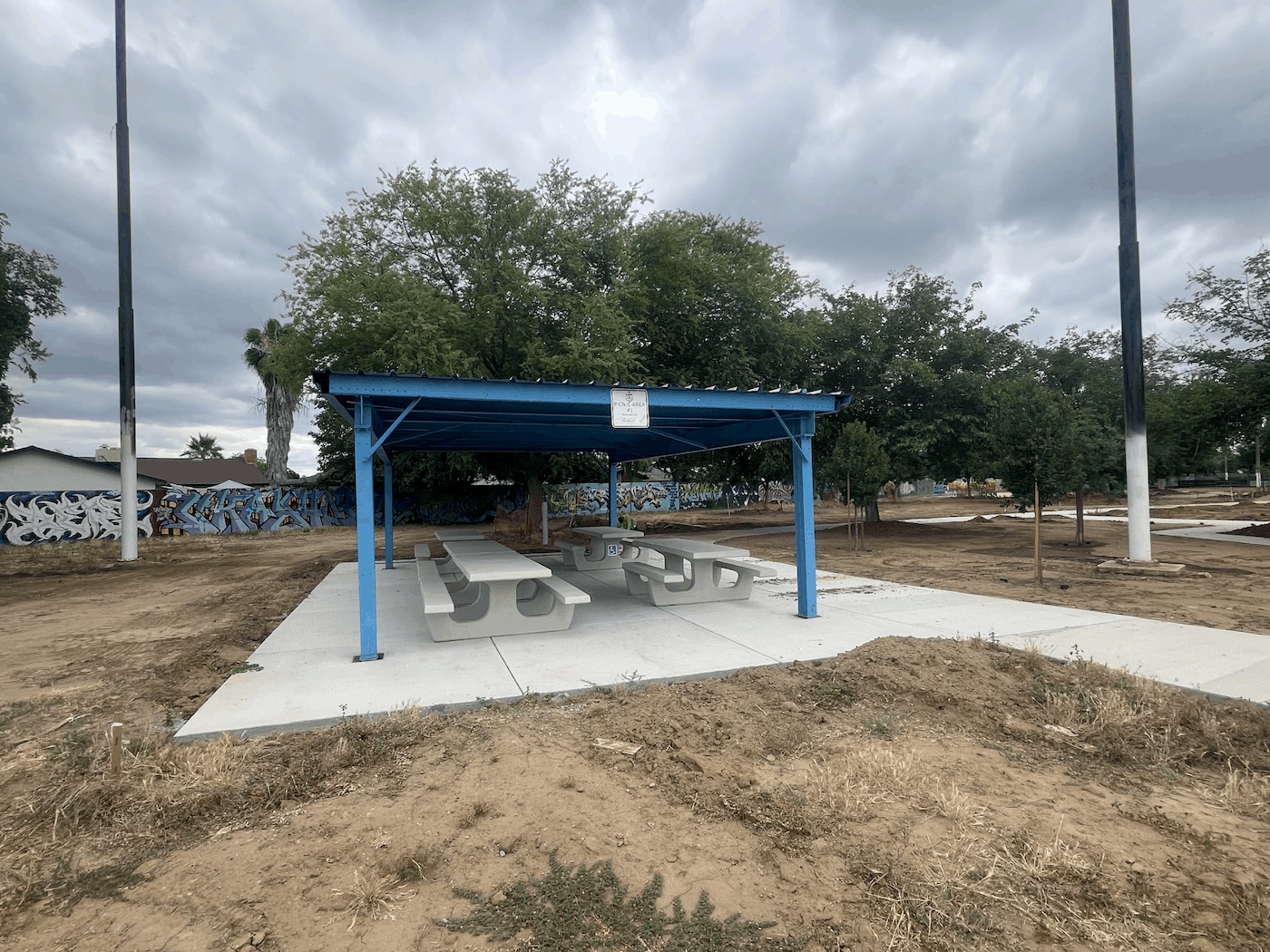 A newly constructed outdoor picnic area featuring a blue metal canopy sheltering several light gray concrete picnic tables. The structure is built on a fresh concrete pad surrounded by bare, graded dirt. In the background, there is a wall covered in graffiti art and several mature trees under a cloudy sky.
