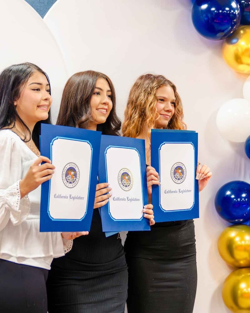 Three young women smile while holding up blue recognition certificates from the California Legislature in front of a balloon decoration.