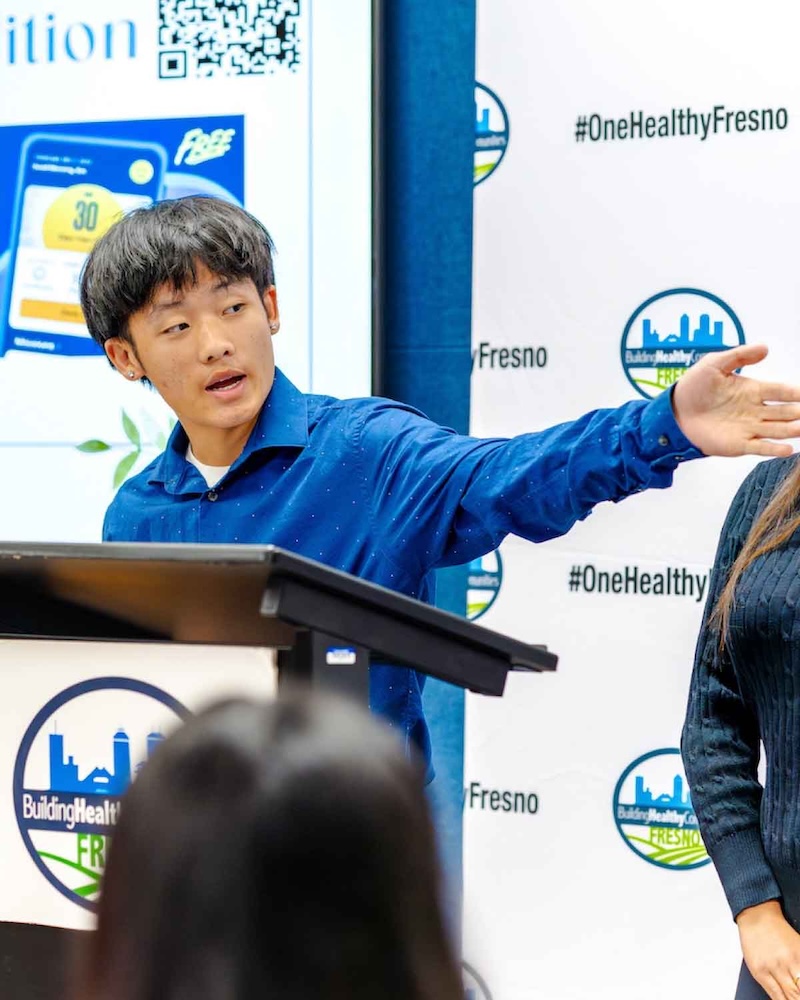 A young man in a blue button-down shirt speaks and gestures with his arm while standing at a podium in front of a #OneHealthyFresno backdrop.