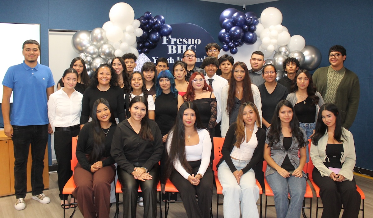 A large group of young adults poses indoors for a group photo in front of a blue and white balloon arch and a sign that reads 