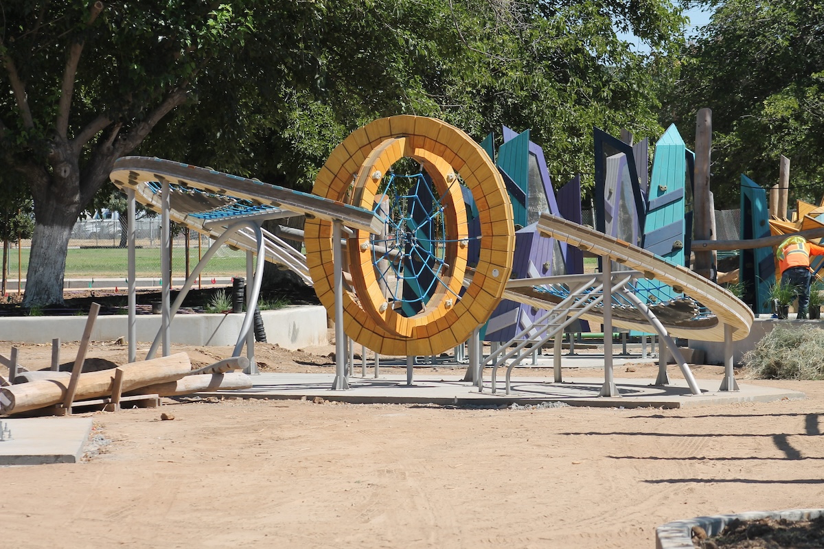 A playground climbing structure under construction featuring a large yellow wooden ring with a blue rope web in the center, flanked by angled climbing nets on metal frames, with tall blue and purple decorative panels in the background.