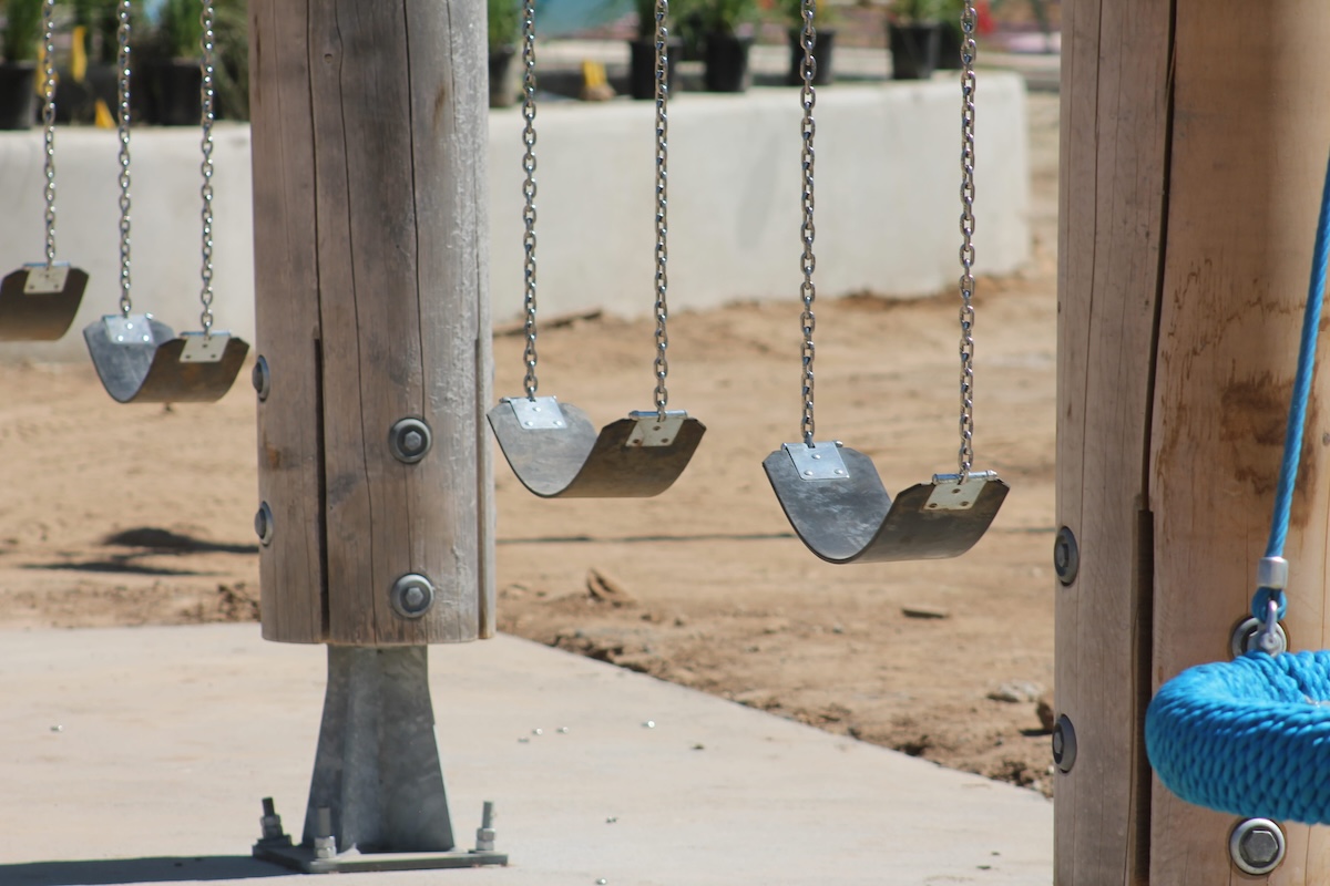 A row of standard black belt swings hangs from metal chains attached to thick cylindrical wooden posts, with a portion of a blue rope nest swing visible on the right edge of the frame.
