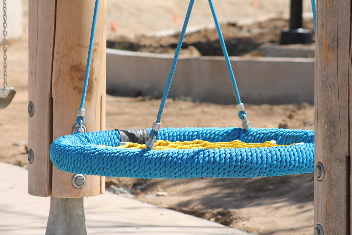 A close-up view of a large, circular nest swing featuring a thick blue braided outer ring and a yellow center, suspended by ropes between sturdy wooden A-frame posts over a concrete pad.