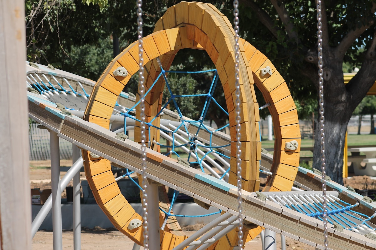  complex climbing apparatus featuring large, intersecting yellow wooden rings filled with blue rope spiderwebs, connected to angled metal frames and climbing nets.