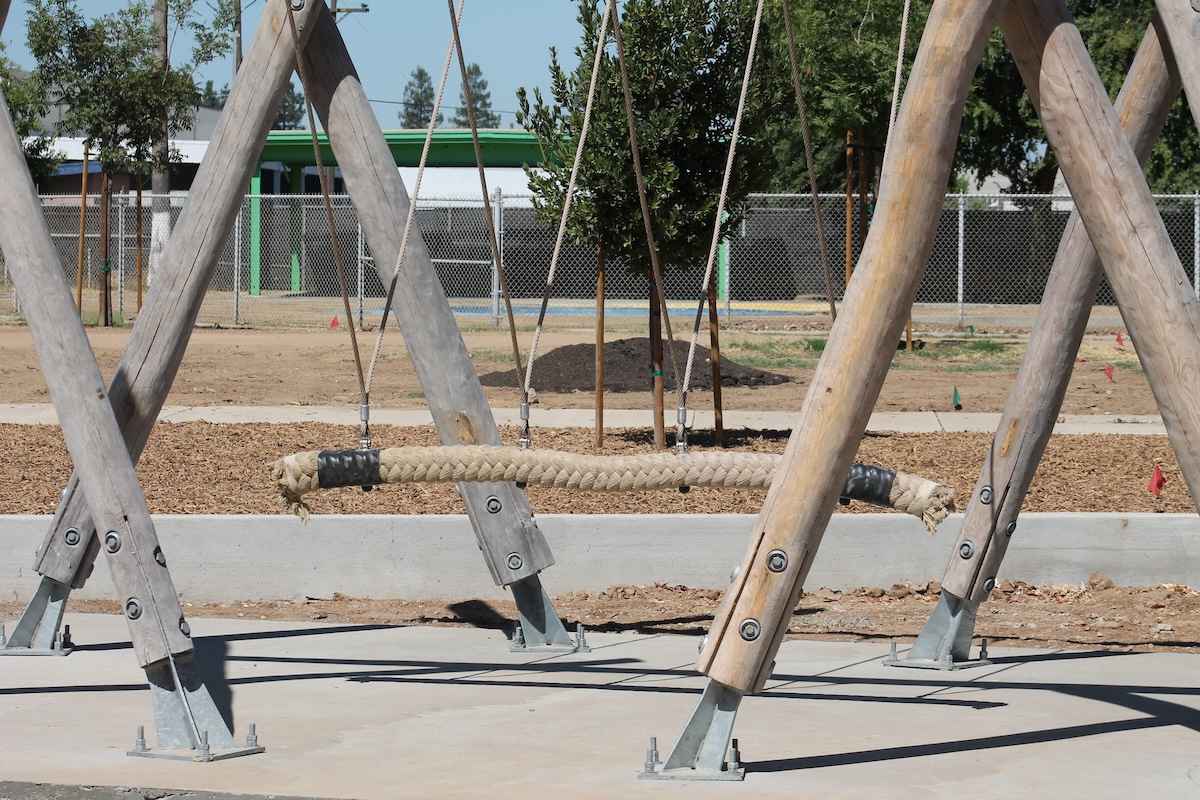 A thick, horizontal rope swing attached to sturdy wooden A-frame supports that are anchored to a concrete pathway.