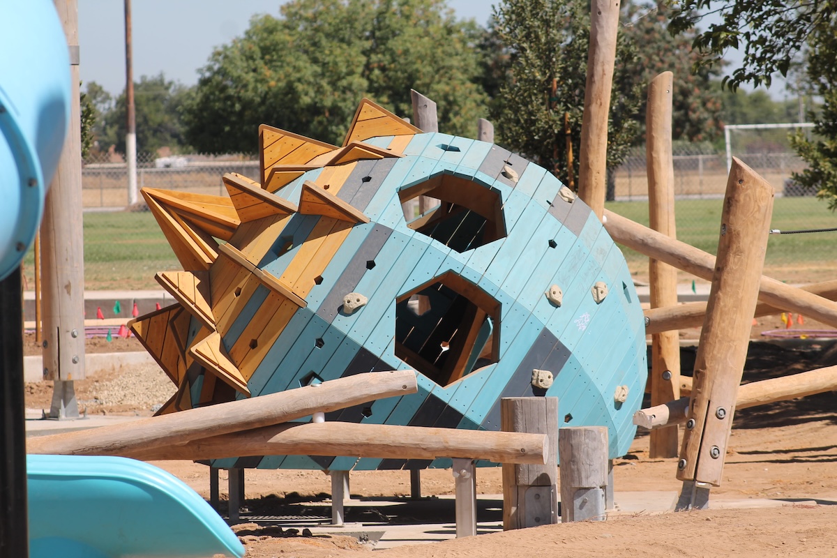 A spherical wooden playground climbing structure painted light blue and yellow, featuring climbing holds, hexagonal windows, and decorative spiky fins on one side.