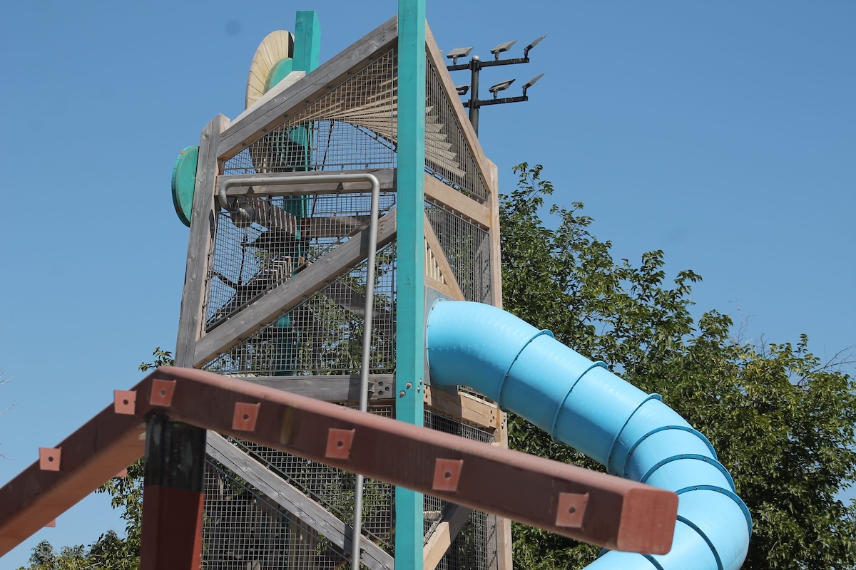 An upward view of a tall, mesh-enclosed wooden playground tower with a blue tube slide extending outward, set against a clear blue sky.