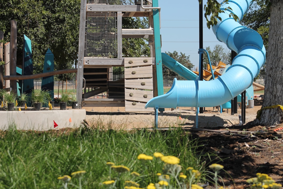 A ground-level view past green grass and yellow wildflowers toward a tall wooden playground tower equipped with a climbing wall and a spiraling blue tube slide.