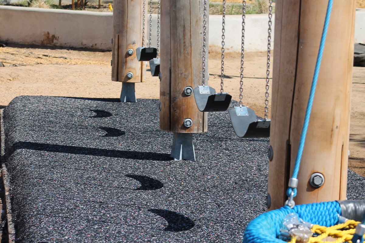 The lower section of a wooden swing set showing black belt swings hanging over a textured, black rubberized safety surface.