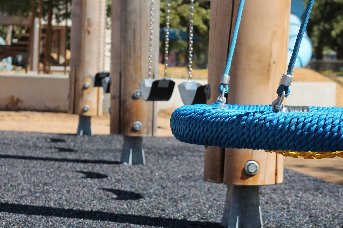 The thick, braided blue rope edge of a circular nest swing is shown in focus in the foreground, while standard black belt swings hanging from wooden posts are visible in the blurred background over a black rubber safety surface.