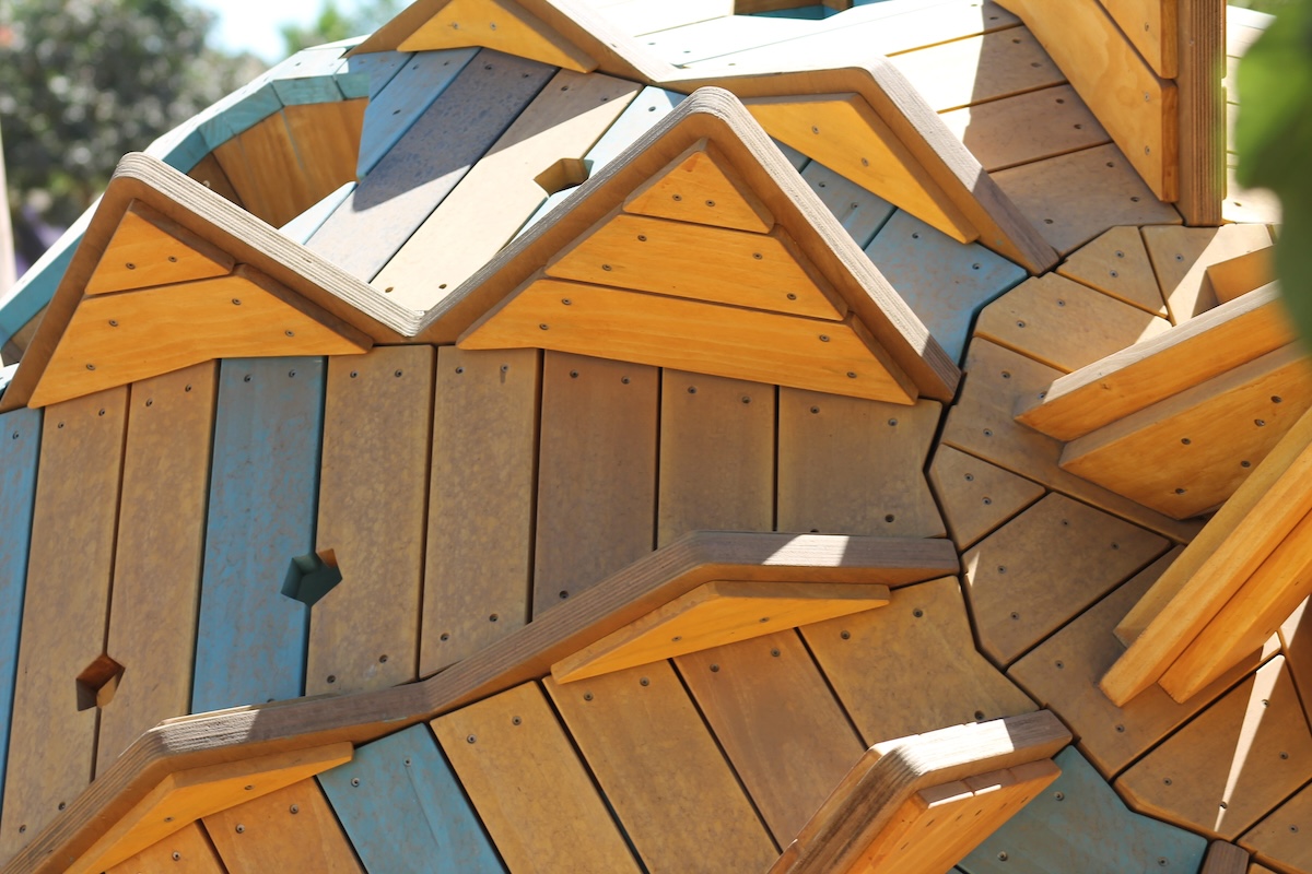 A detailed close-up of a climbing structure built from interlocking natural wood and light blue panels, showing triangular ridges and small hexagonal cutouts used as climbing holds.