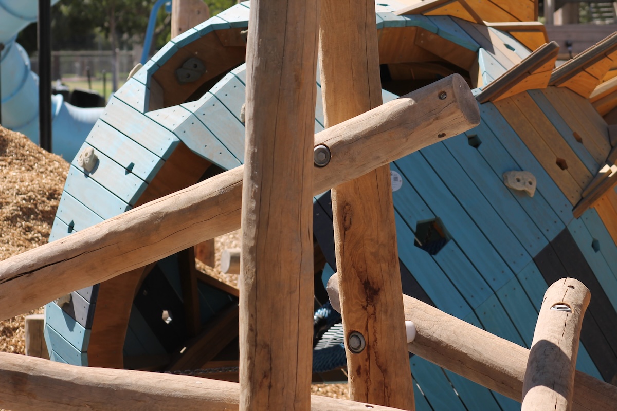 A close-up of a playground structure showing thick wooden support beams in front of a light blue rounded climbing feature with hexagonal cutouts and small climbing holds.
