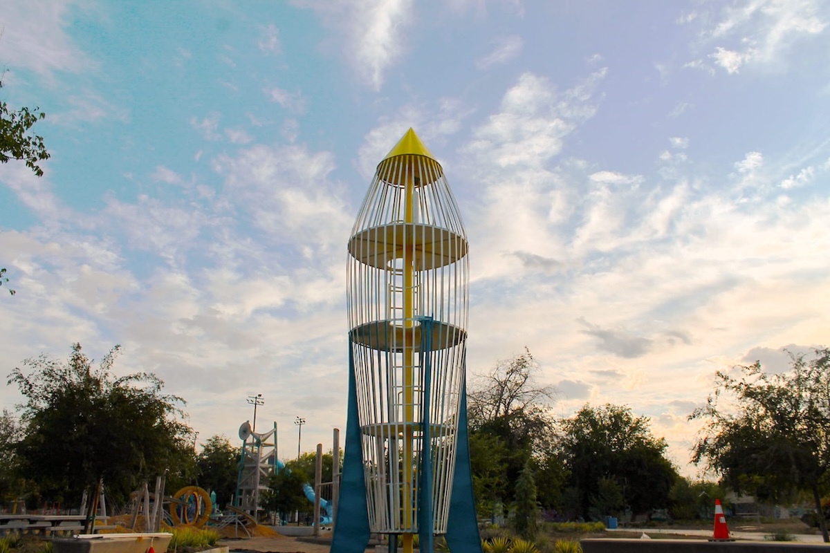 A tall, retro-style rocket ship playground climbing structure made of vertical white metal bars with a yellow cone top and blue base fins, featuring multiple internal platforms accessible by a central ladder against a cloudy sky.