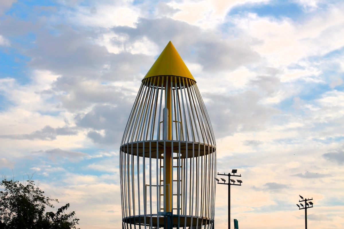 The upper section of a tall playground structure designed to look like a retro rocket ship, featuring a yellow conical roof, white vertical metal bars forming the hull, and an internal ladder, all set against a partly cloudy sky.